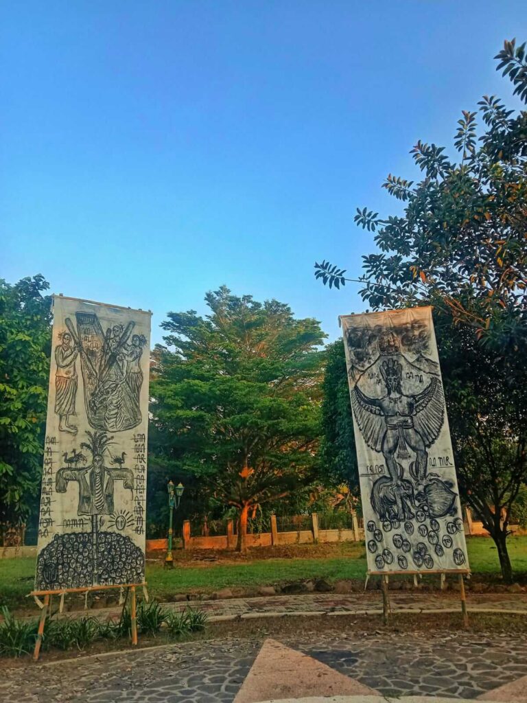 Two vertical banners in a park each with large black ink drawings of mythic figures and symbols against a clear blue sky and green trees behind